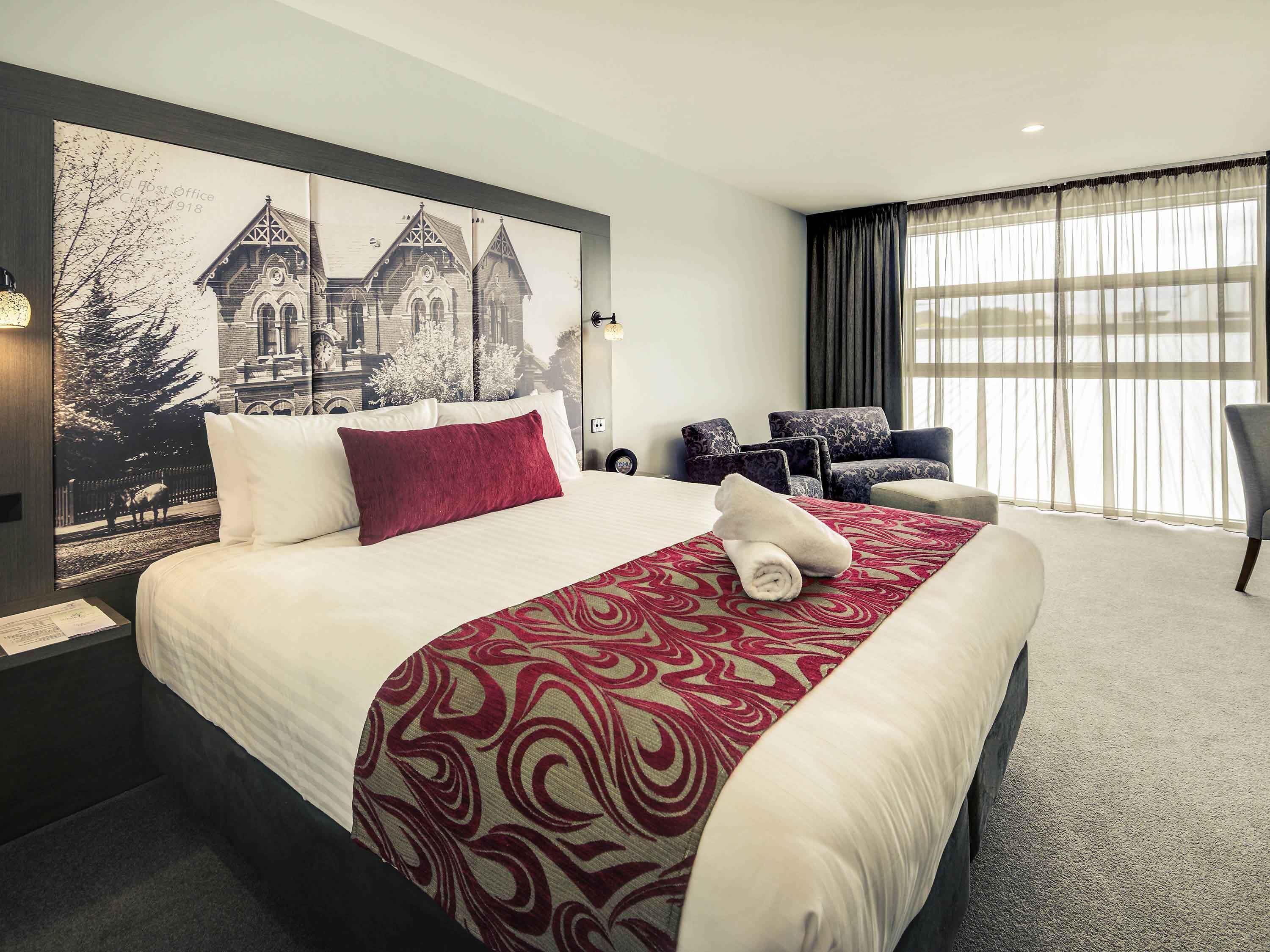 A hotel room with a large bed, a headboard with a cityscape photo, a seating area with chairs, and a large window with curtains.