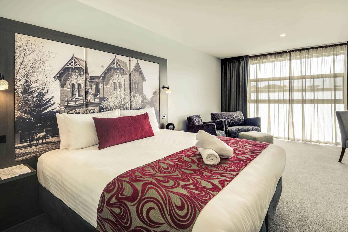 Una habitación de hotel con una cama grande, un cabecero con una fotografía de un paisaje urbano, una zona de estar con sillas y un ventanal con cortinas.