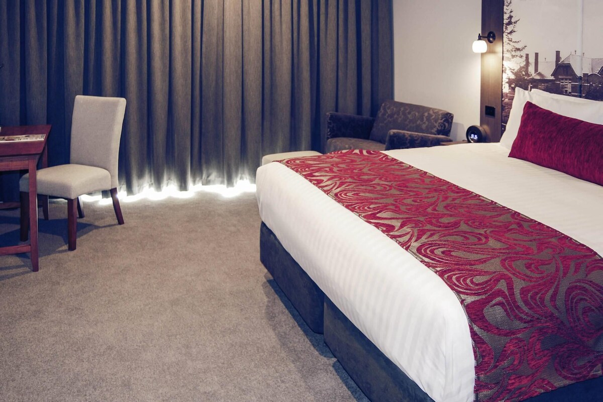 A hotel room with a large bed, a desk, a chair, and a view of a building through the window.
