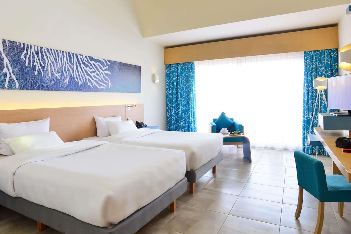 A hotel room with two beds, a desk with a computer, a chair, and a large window with curtains.