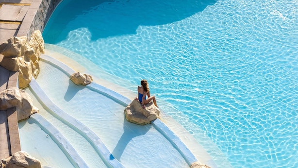 A pool with clear water and rocks, a person sitting on a rock, and a tiled deck. - Novotel Marsa Alam Beach Resort (El Quseir)