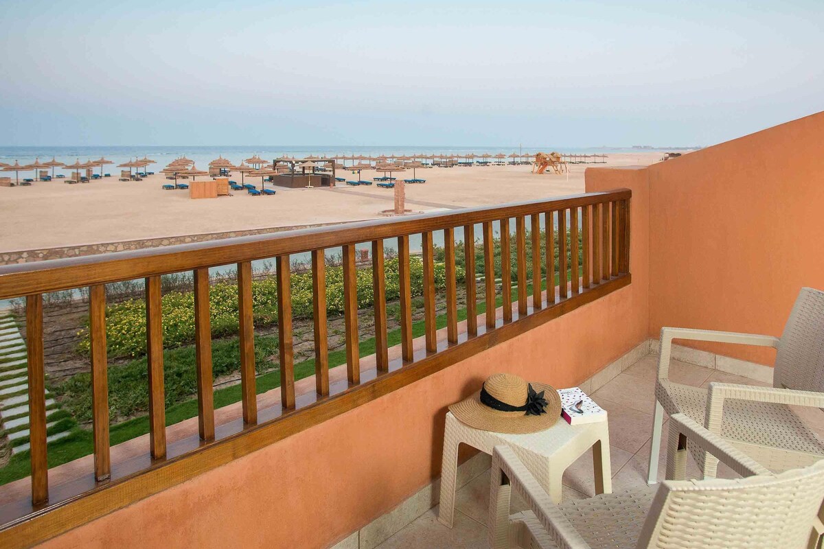 A balcony with a view of a beach, a small table with a hat, and two wicker chairs.
