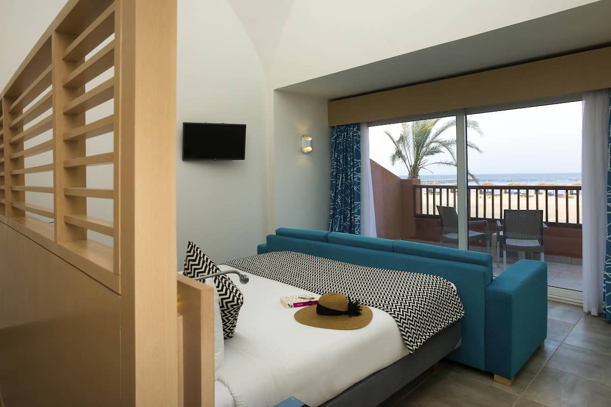 A room with a bed, a TV, a balcony with a view of the beach, and a window with curtains.
