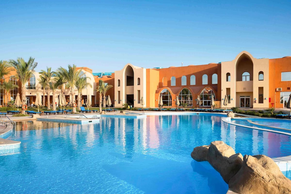 A hotel with a large swimming pool, palm trees, and a modern building with arched windows.