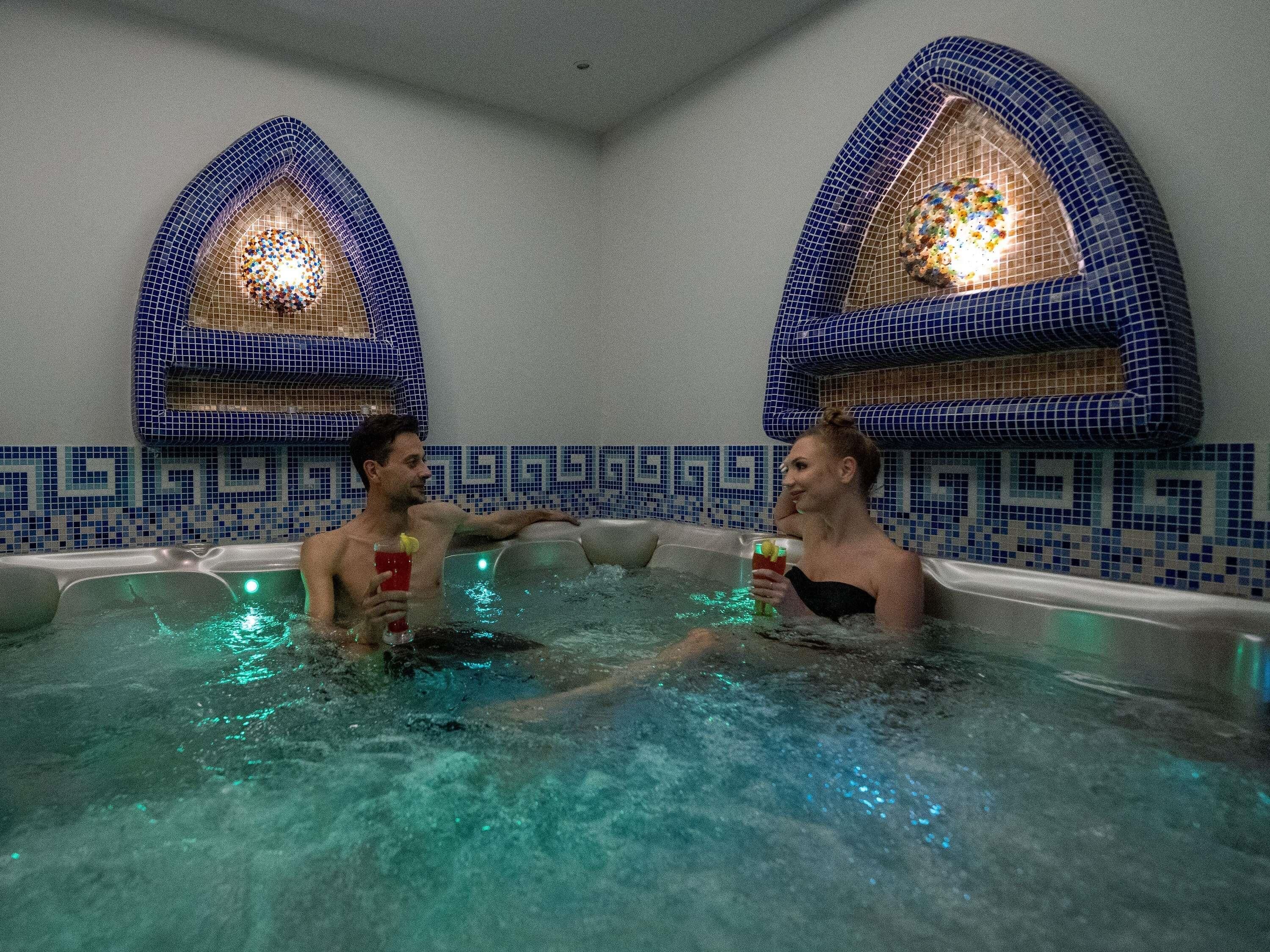 Two people in a hot tub with tiled walls and arched windows.