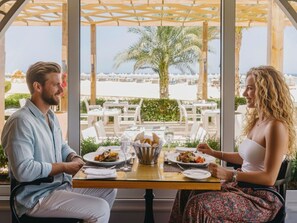 A dining area with tables and chairs, a view of the beach, and palm trees. - Novotel Marsa Alam Beach Resort (El Quseir)