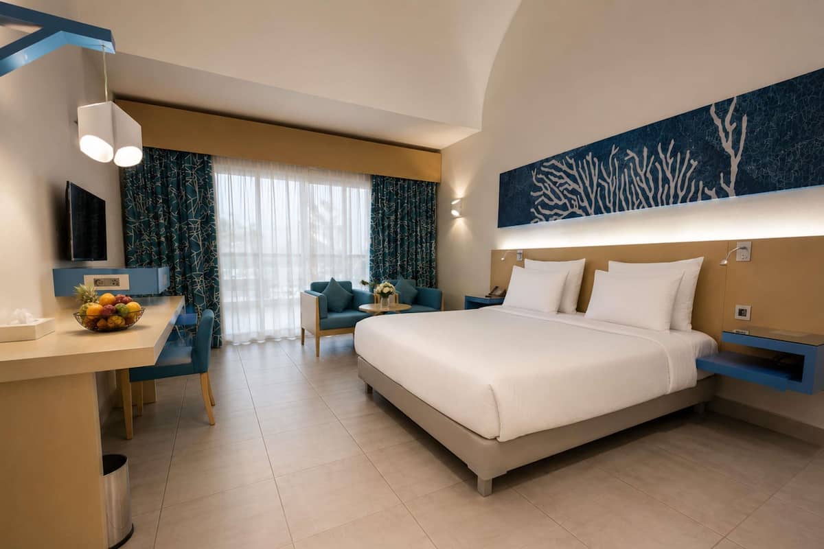 A hotel room with a bed, a desk with a fruit bowl, a chair, a TV, and a wall art piece.