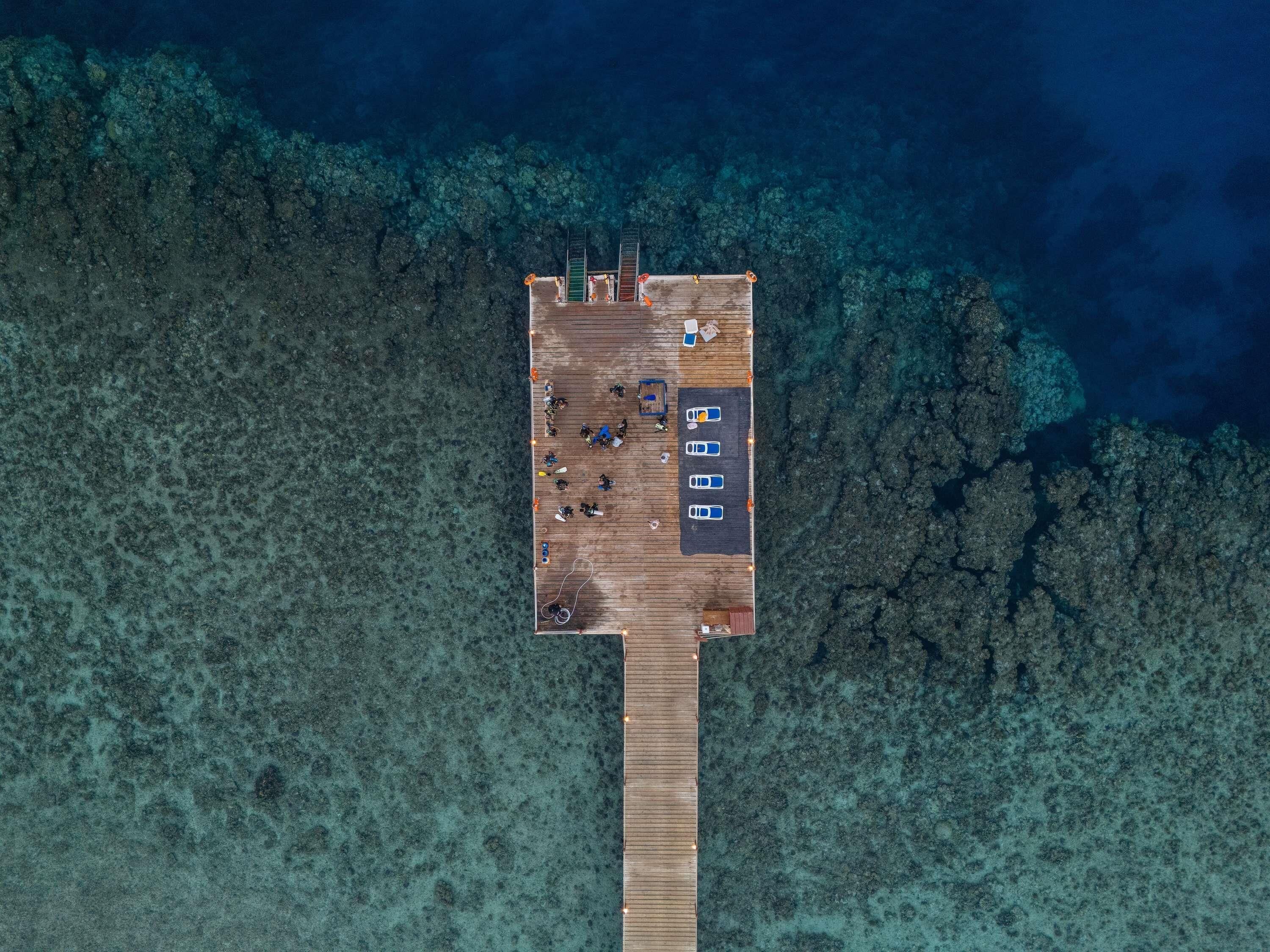 An aerial view of a wooden pier extending into clear blue water, with a parking area and a few people on the pier.