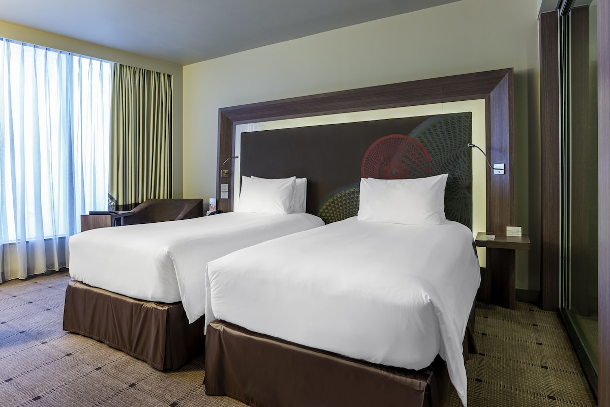 Een hotelkamer met twee bedden, een bureau, een stoel en een groot raam met gordijnen.