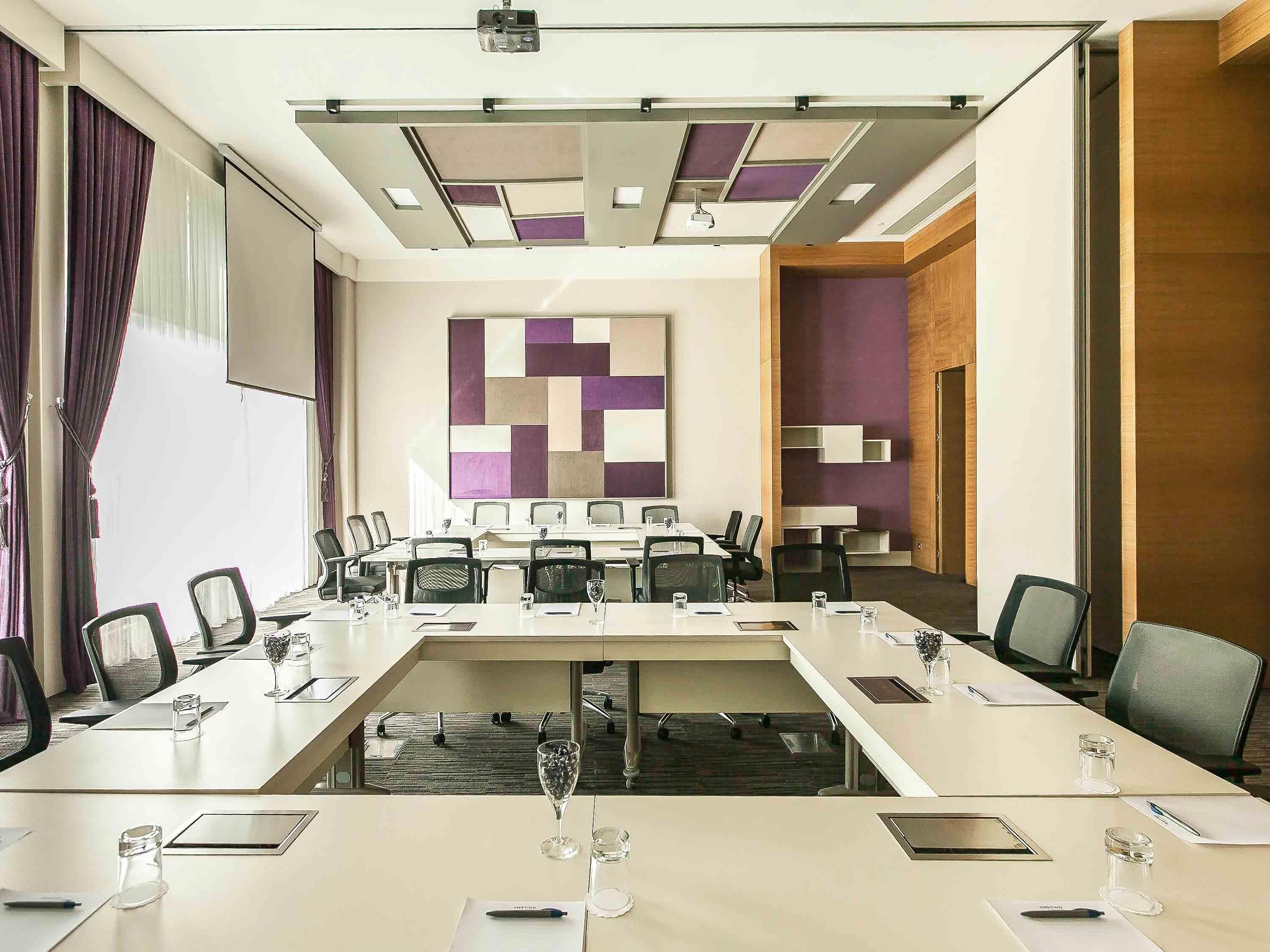 Un salón de conferencias con una mesa larga, sillas, un proyector y una gran decoración abstracta en la pared.