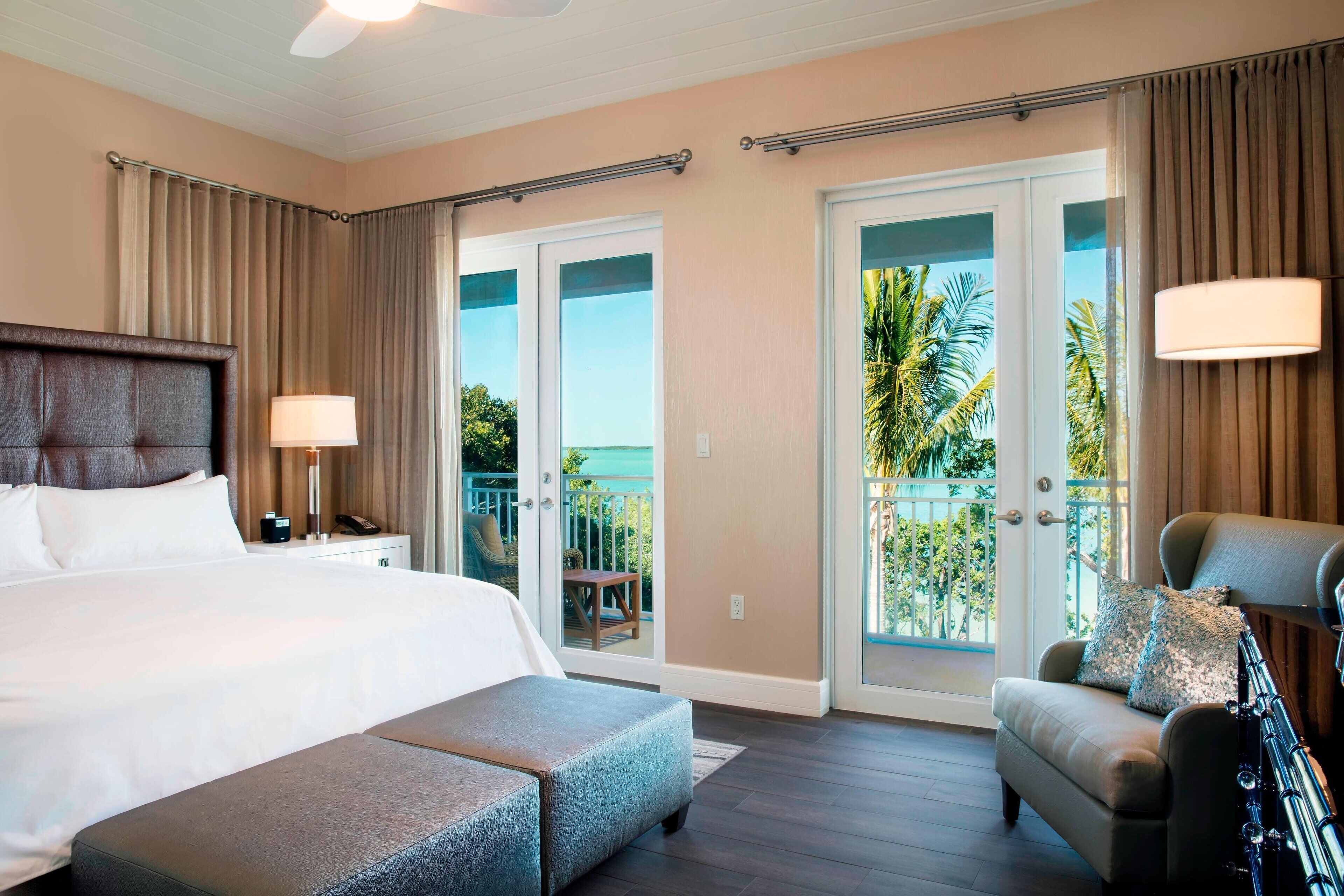 A hotel room with a large bed, a seating area with a chair and ottoman, a balcony with a view of palm trees, and a ceiling fan.