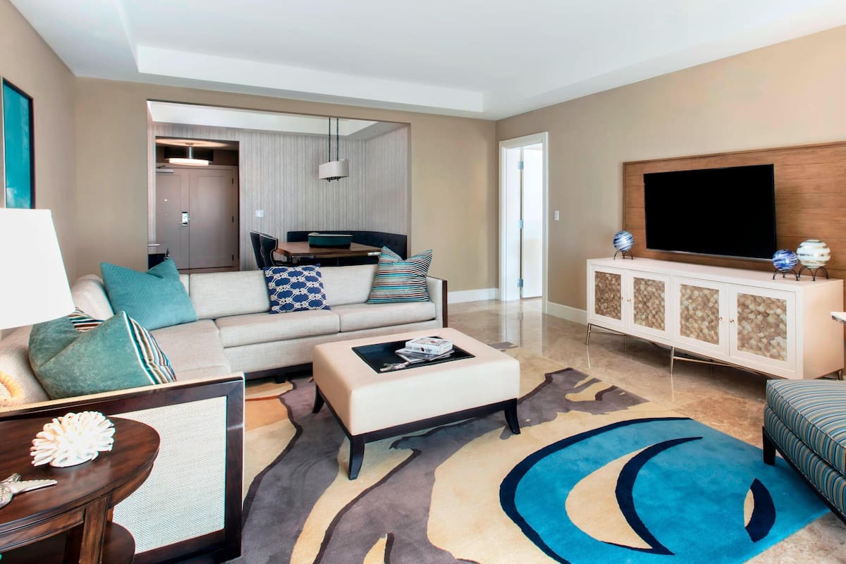 A modern living room with a sofa, ottoman, and a flat-screen TV on a cabinet. There is a dining area in the background.