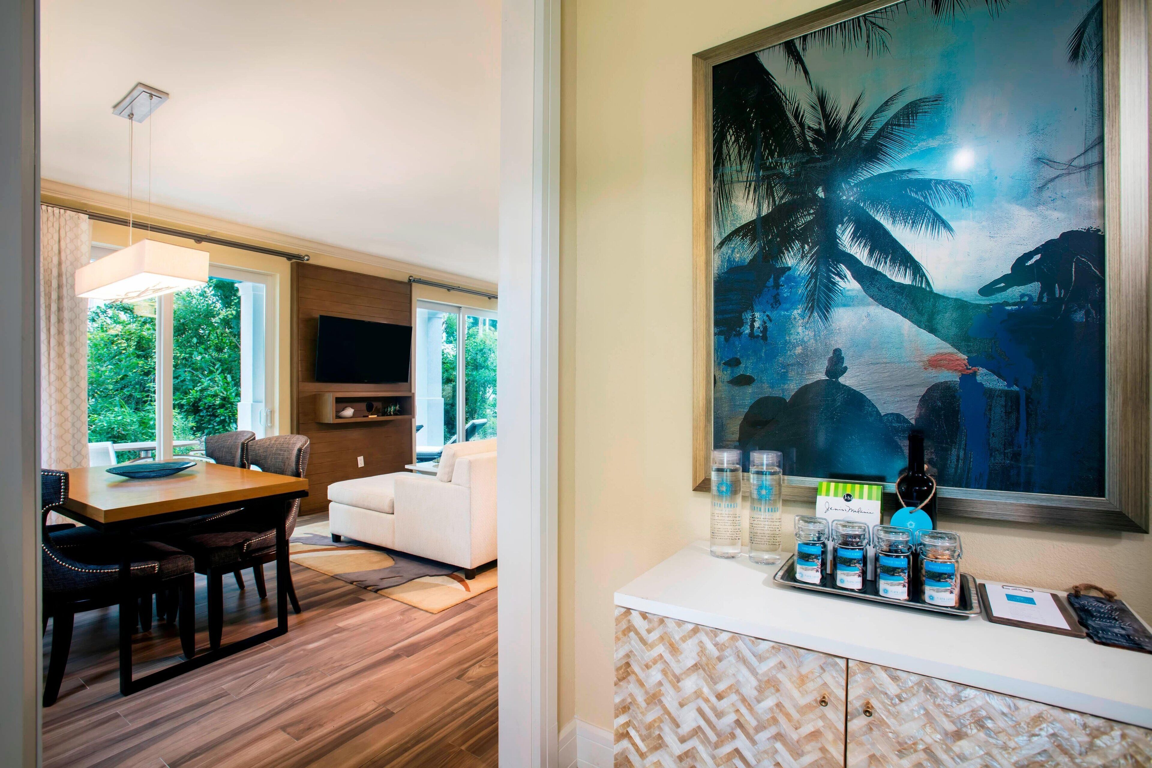 A modern living room with a dining area, large windows, and a painting of a tropical scene.