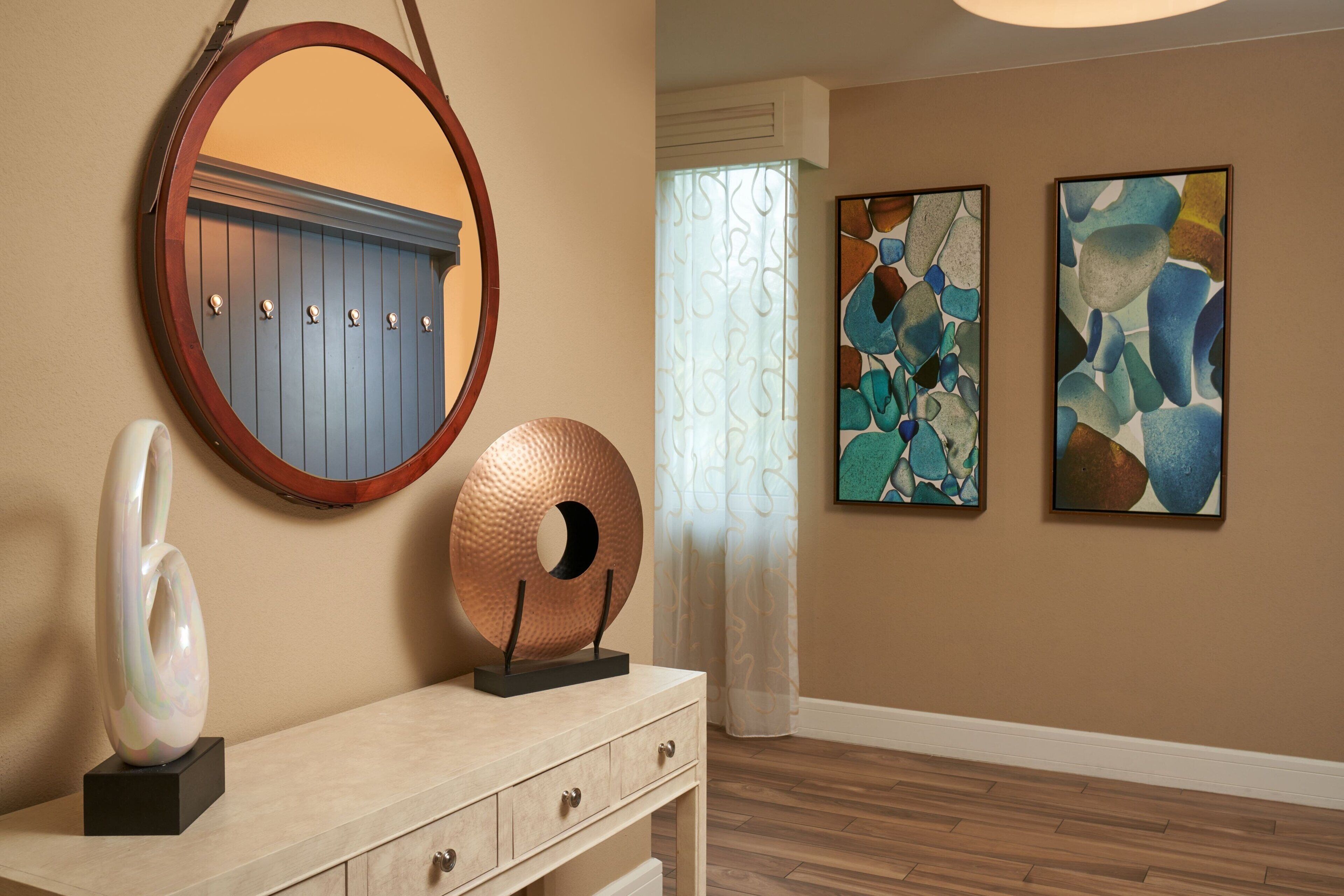 Une pièce dotée d’un plancher en bois, d’un miroir rond, d’une sculpture décorative, d’une commode à tiroirs et de deux tableaux abstraits accrochés au mur.