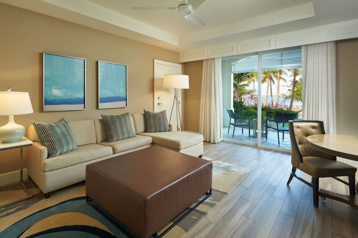A hotel room with a sofa, ottoman, dining table, and chairs. There is a lamp, a floor lamp, and two framed pictures on the wall. The room has a large window with a view of palm trees and a beach.