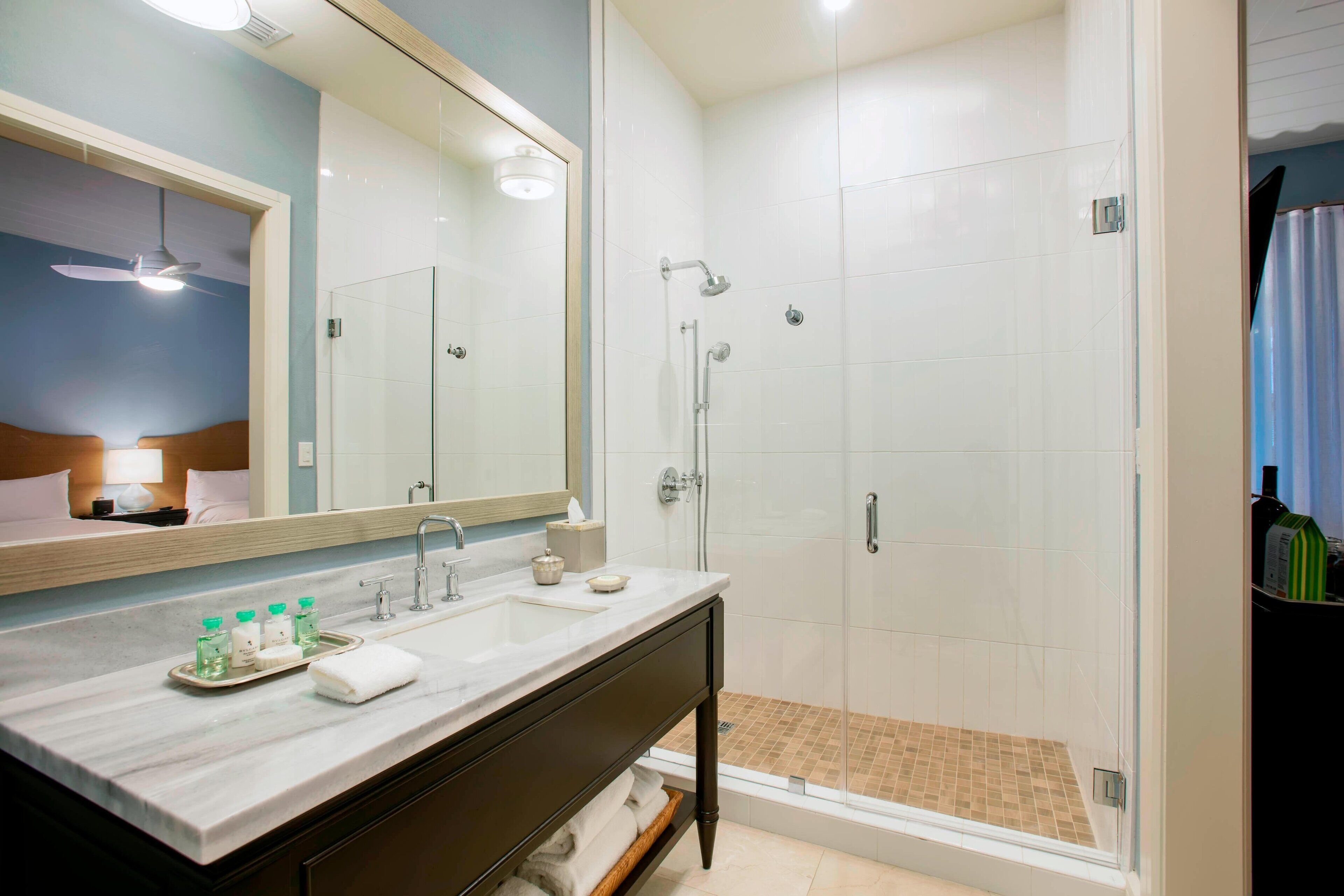 A bathroom with a large mirror, a sink with toiletries, a shower with a glass door, and a towel rack.