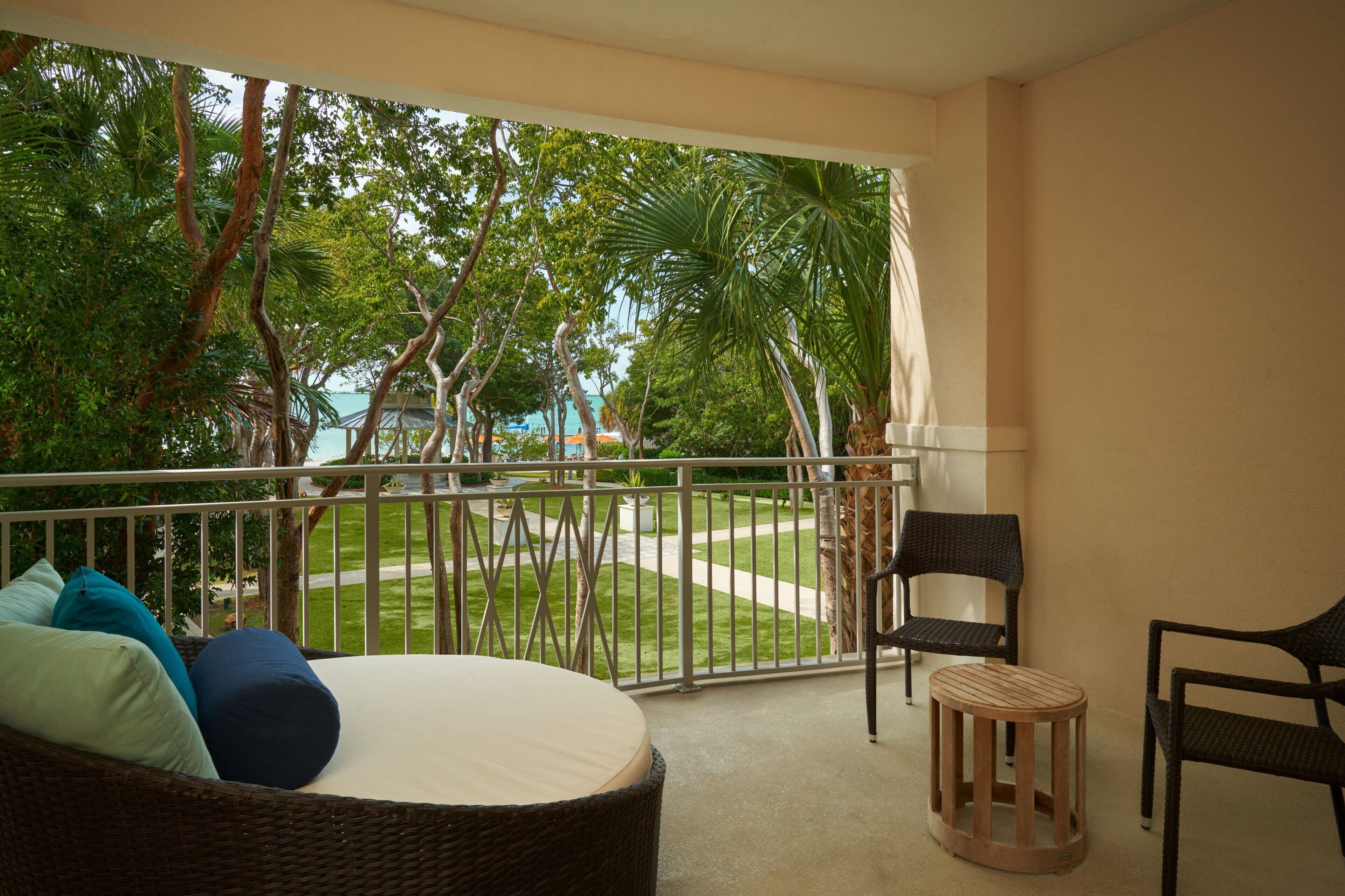 A balcony with wicker furniture, a small round table, and a view of a well-maintained garden and palm trees.
