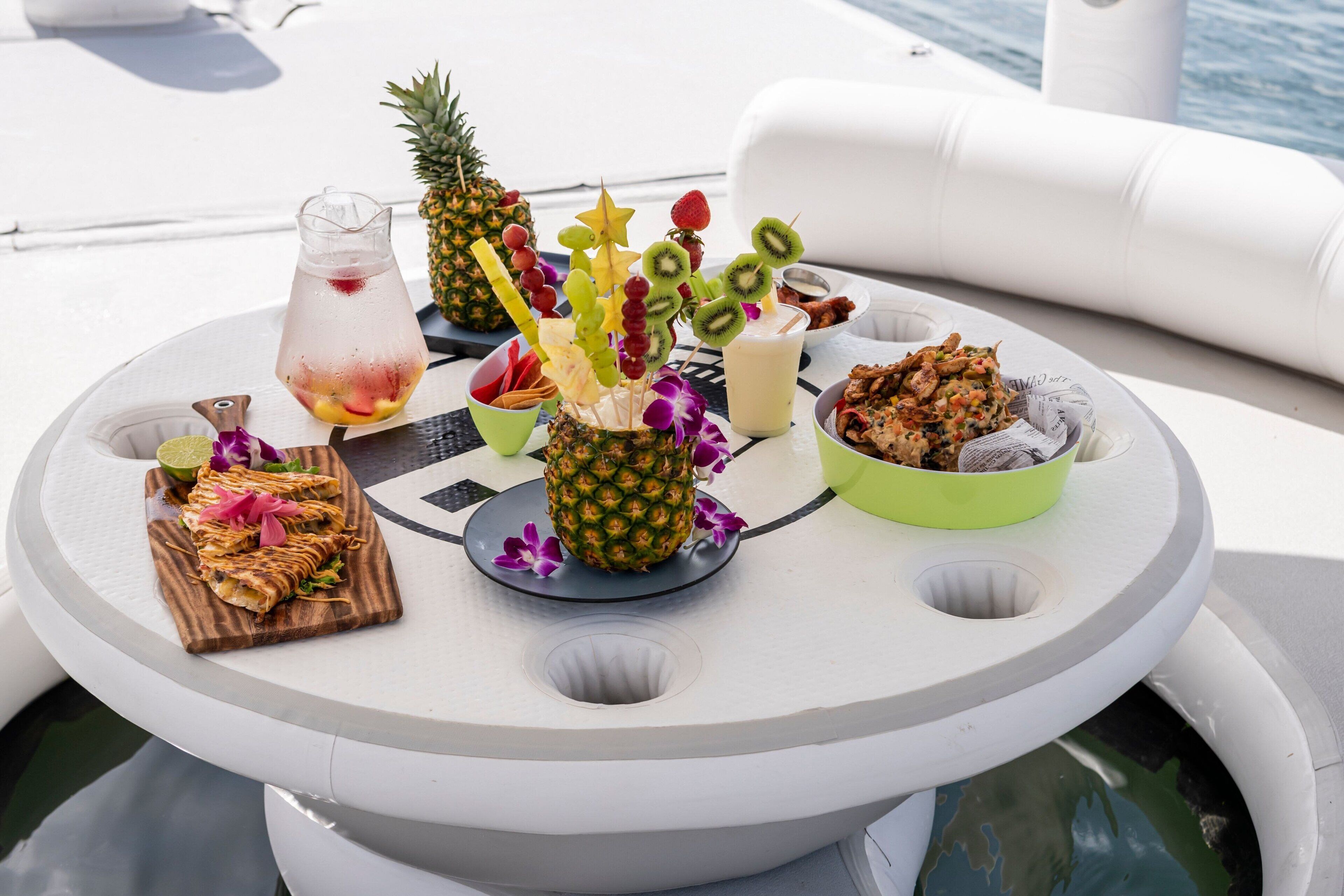 A round table with a variety of food and drinks, including a pineapple, fruit skewers, and a drink pitcher, on a boat deck.