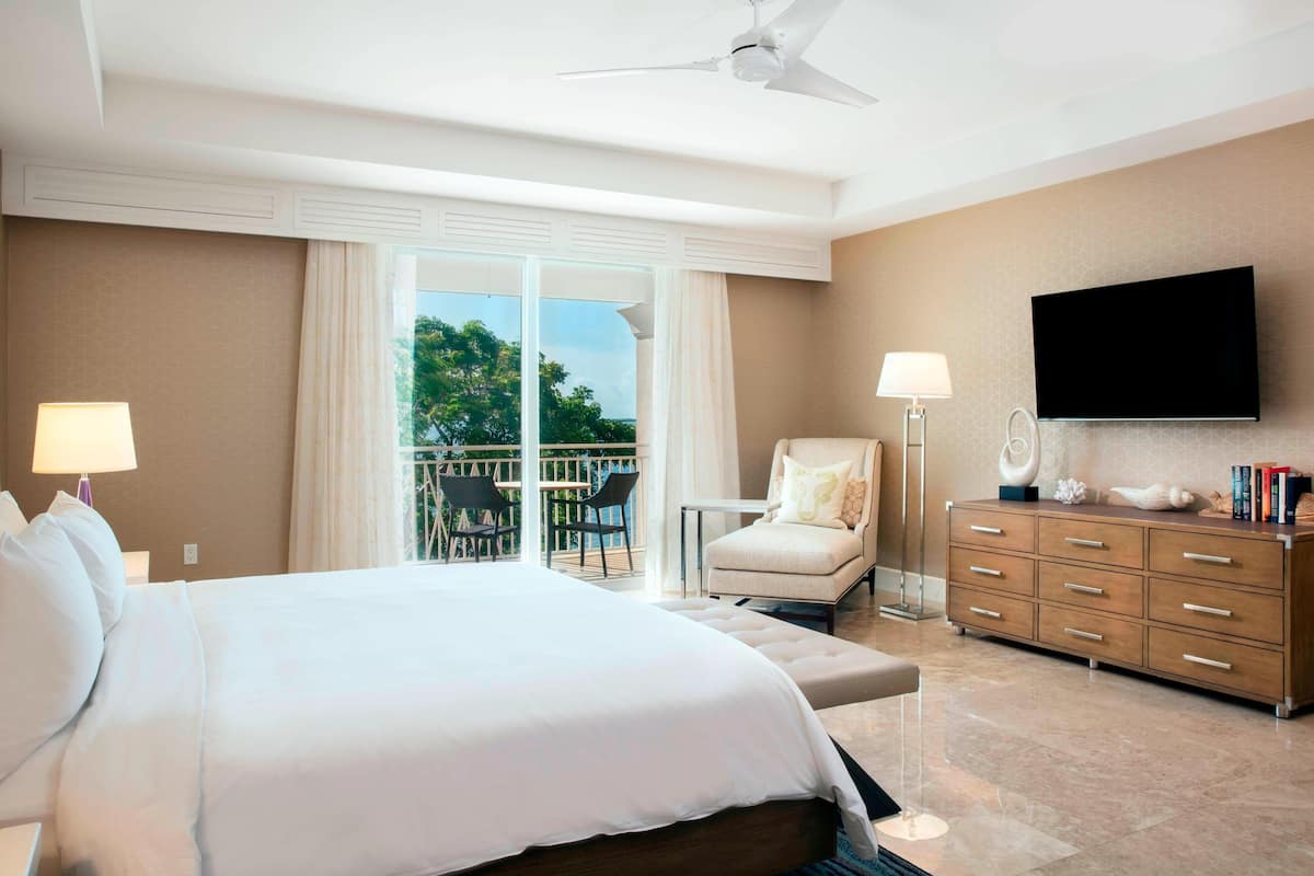 A hotel room with a large bed, a TV mounted on the wall, a chair, a desk, and a balcony with a view of trees.