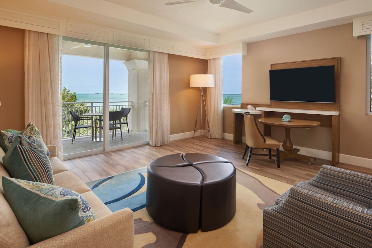 A hotel room with a balcony, a sofa, a round ottoman, a small table, a chair, a lamp, and a TV.