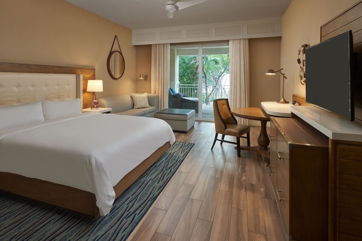 A hotel room with a large bed, a desk, a chair, a sofa, and a balcony with a view of greenery.