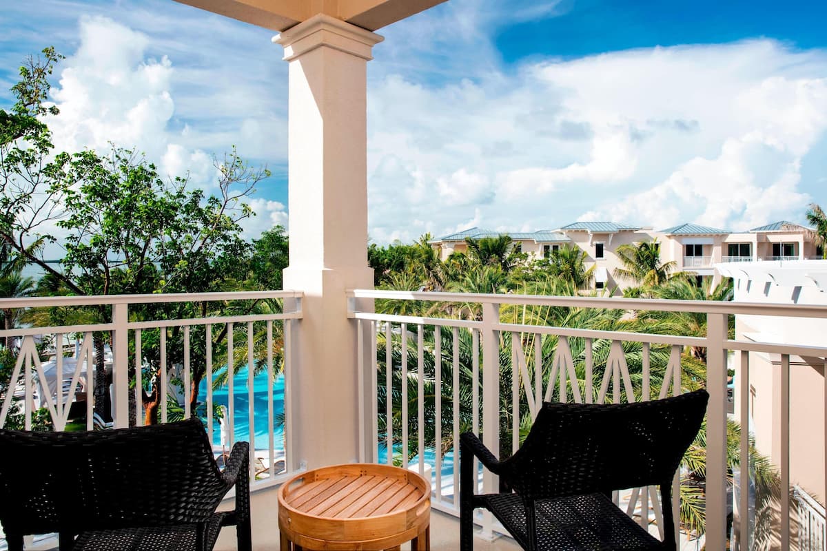 A balcony with wicker chairs and a wooden table, overlooking a swimming pool and lush greenery.