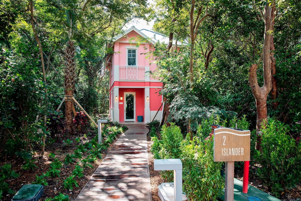 A pink house with a balcony, surrounded by lush greenery and a wooden pathway leading to the entrance.