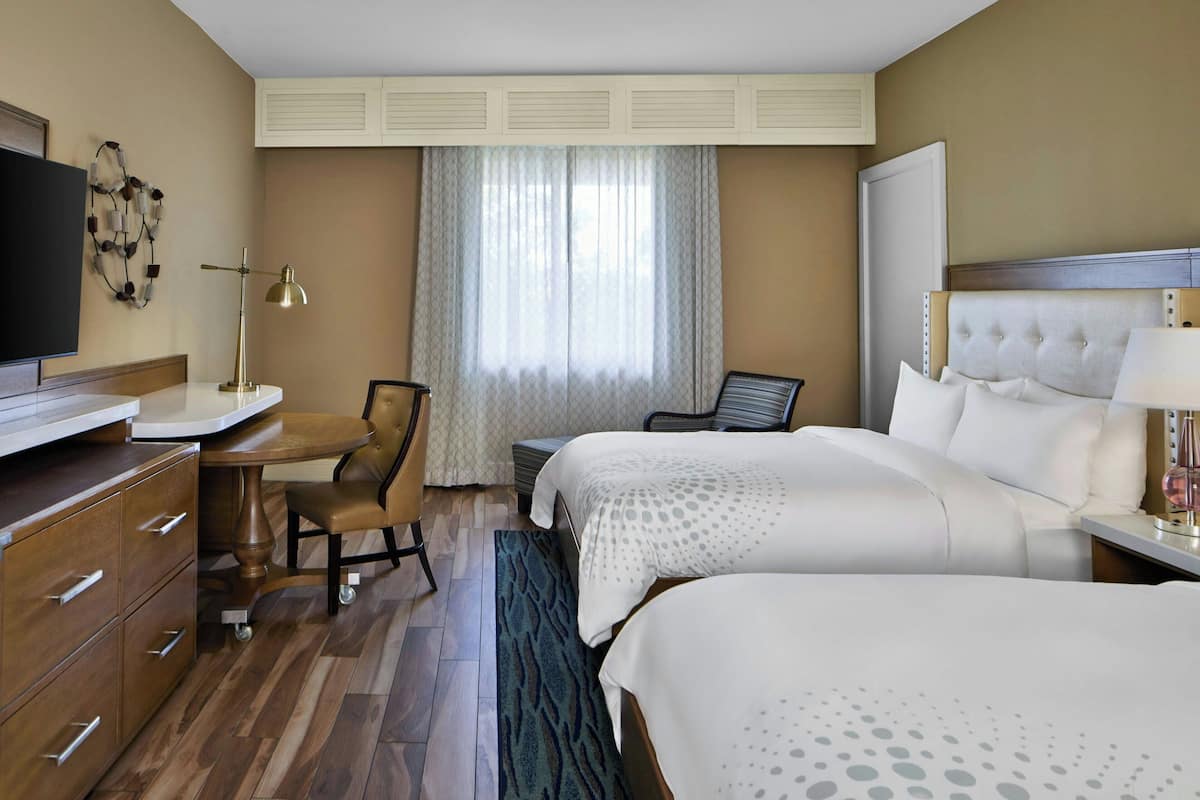 A hotel room with two beds, a desk, a chair, a television, and a window with curtains.