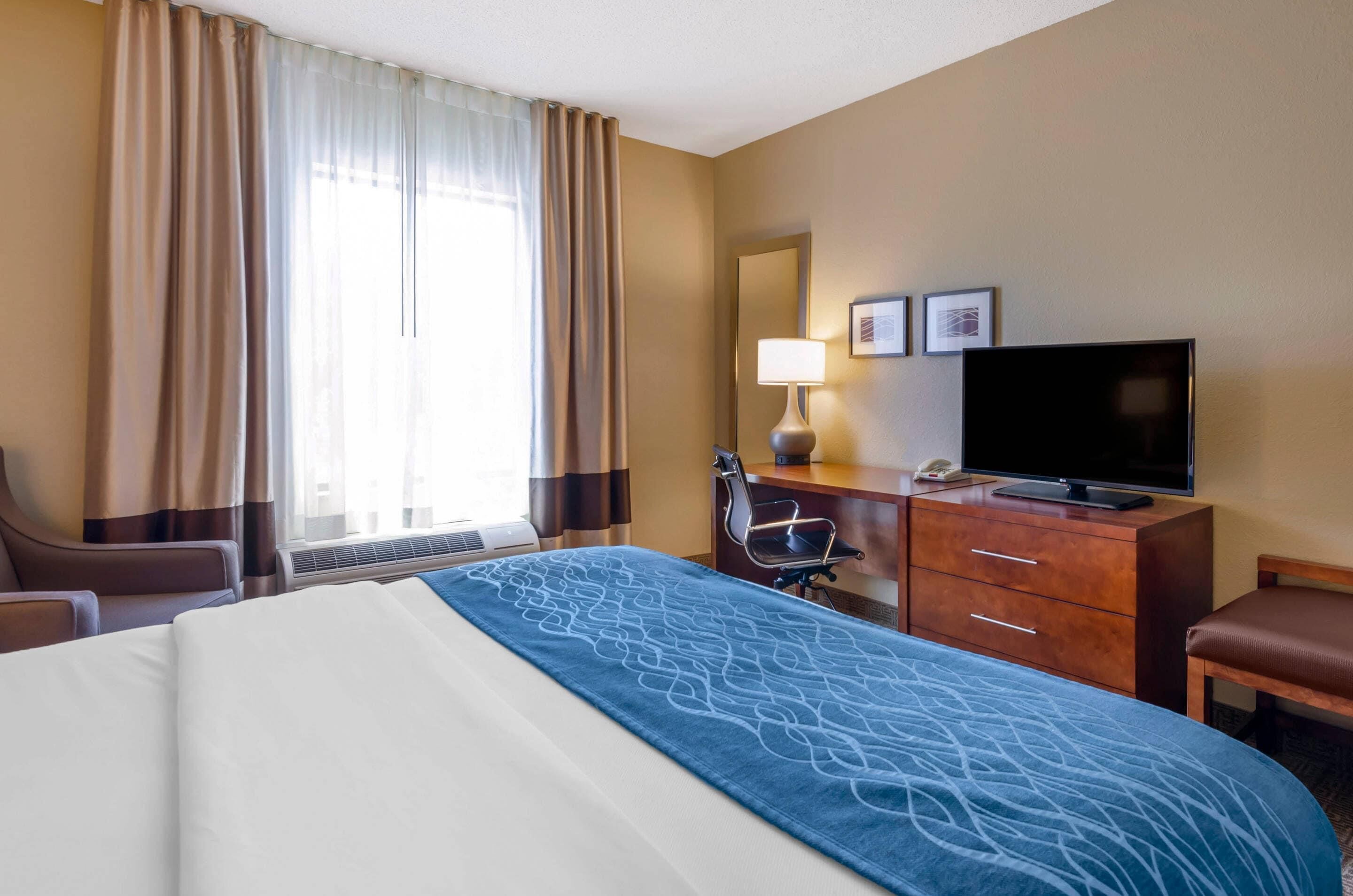 A hotel room with a bed, a desk with a TV, a chair, and a desk lamp.