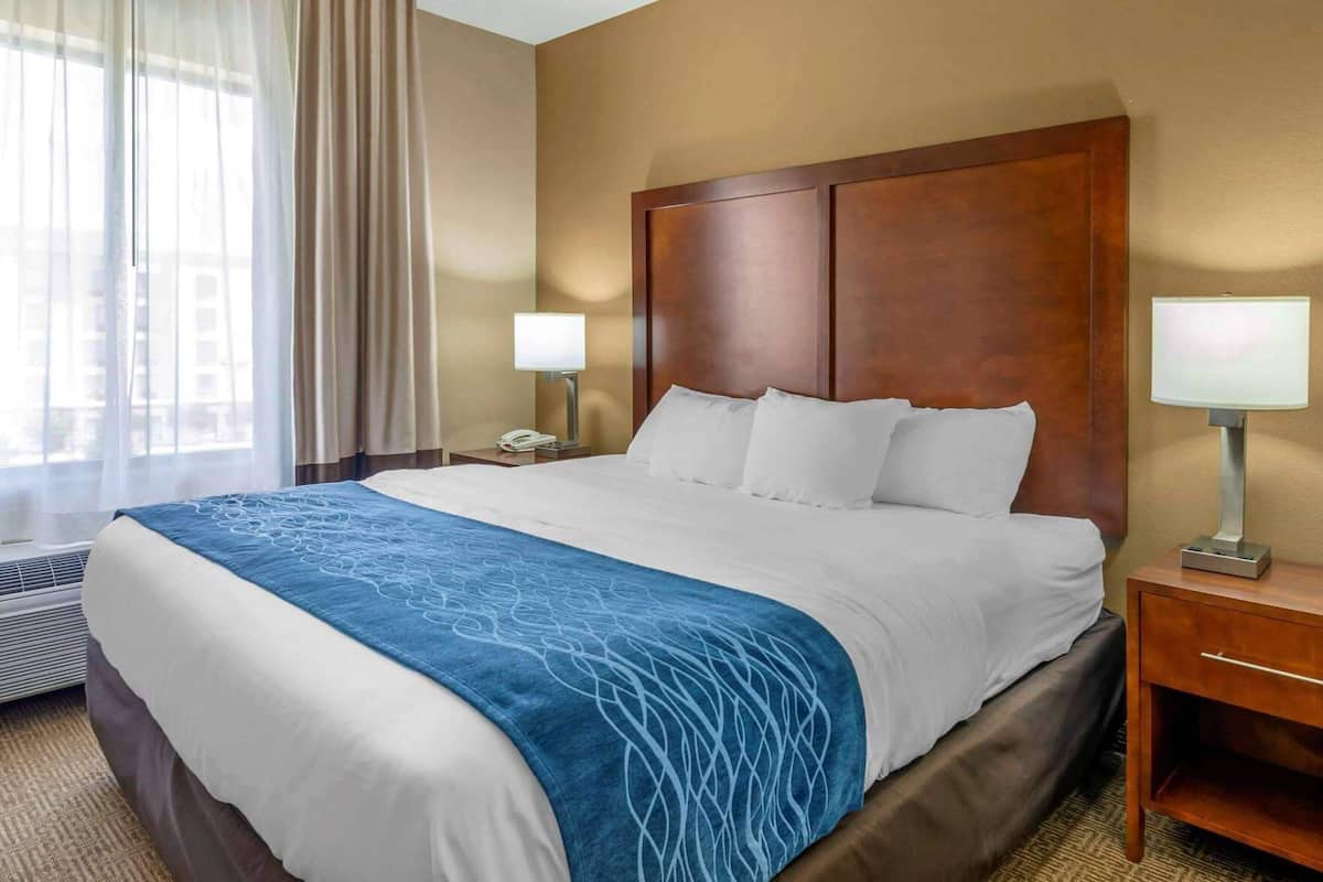 A hotel room with a large bed, a wooden headboard, a bedside table with a lamp, and a window with curtains.