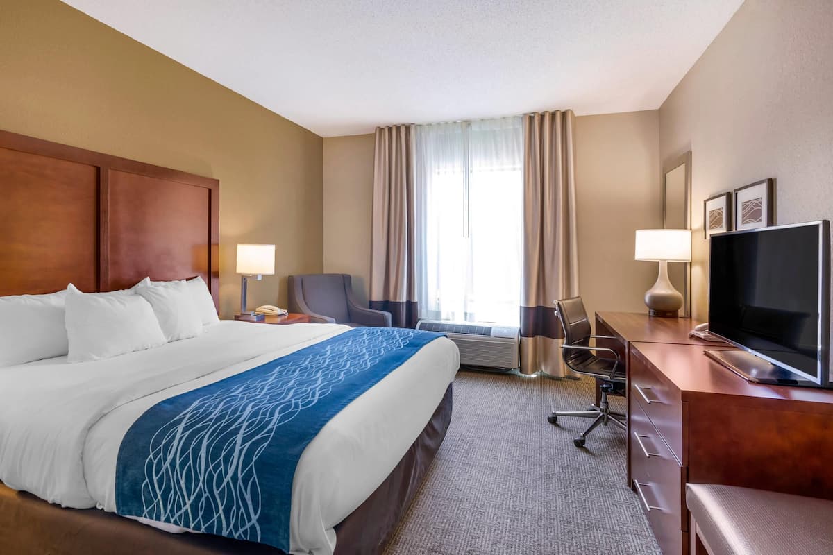 Une chambre d’hôtel avec un grand lit, un bureau avec une télévision, une chaise, une lampe et une fenêtre avec des rideaux.
