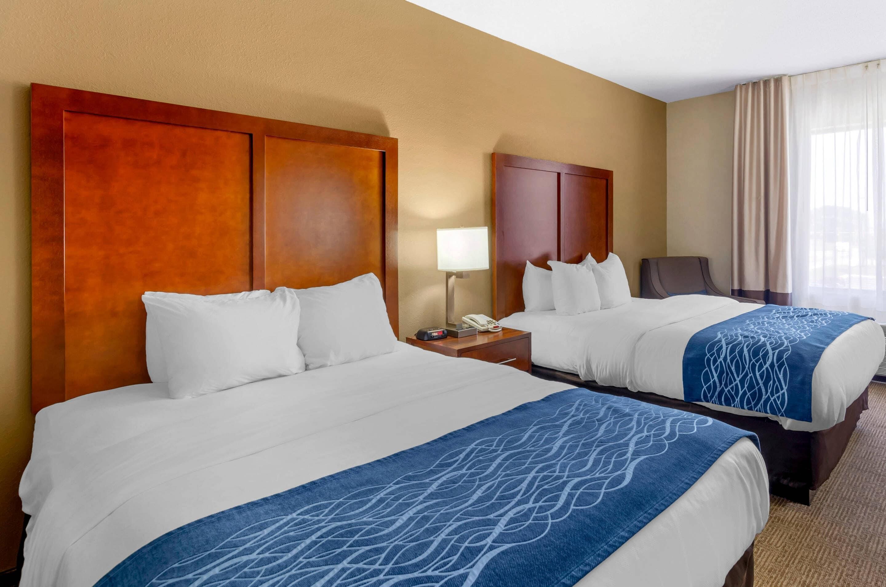 A hotel room with two beds, a nightstand with a lamp, a chair, and a window with curtains.