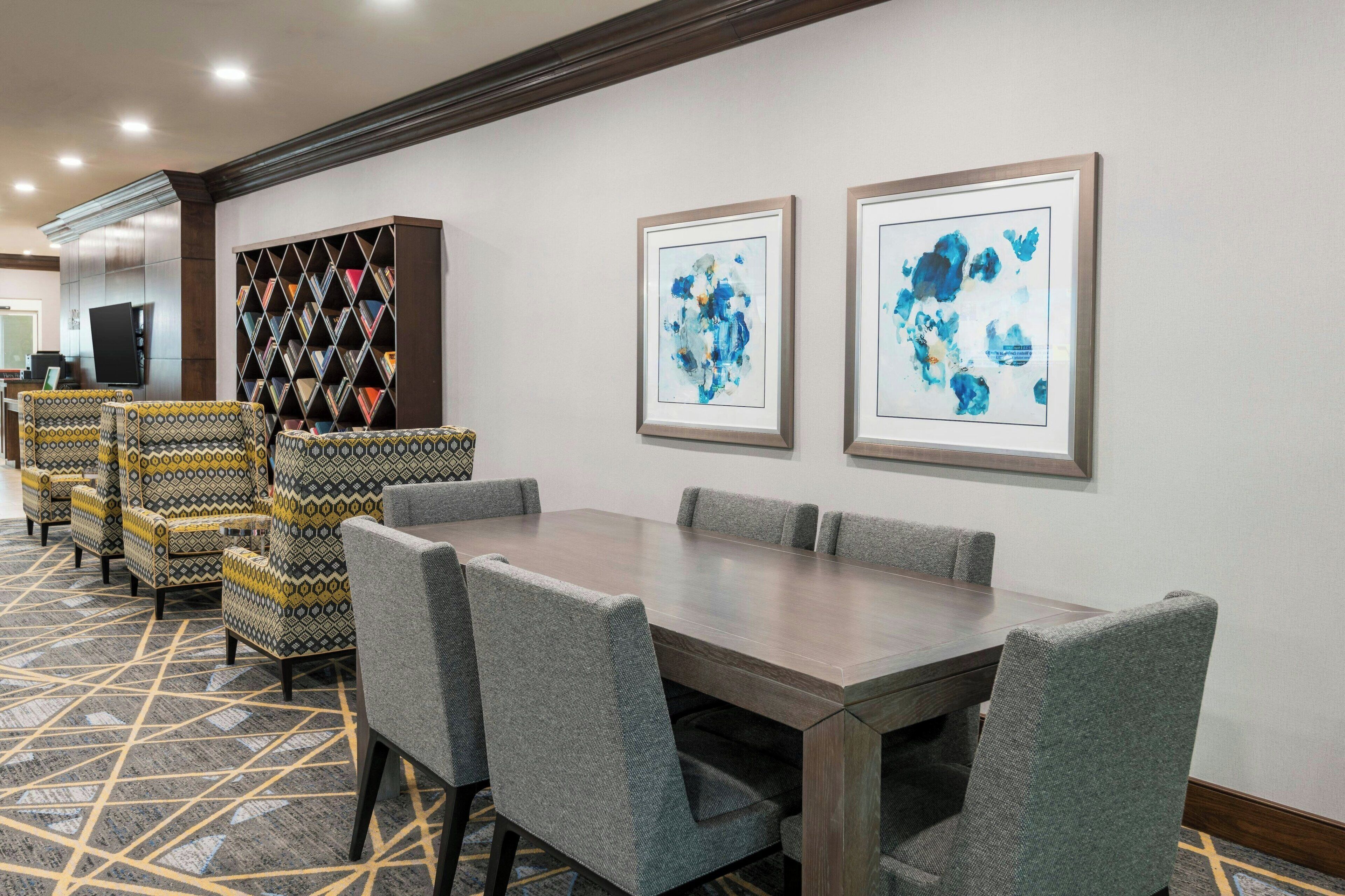 Quarto de hotel com uma grande mesa de jantar, cadeiras estofadas cinzentas, carpete estampado, uma estante de livros e duas pinturas abstratas emolduradas na parede.