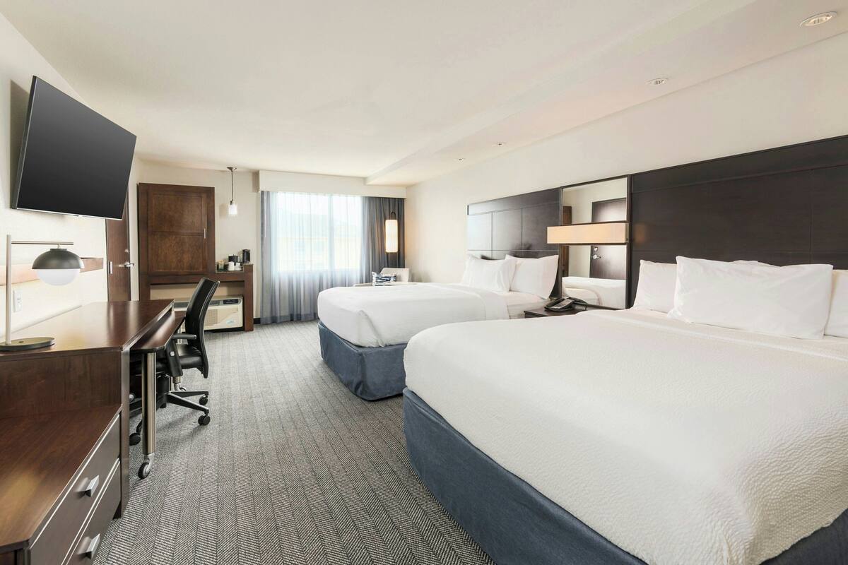 Habitación de hotel con dos camas, un escritorio, una silla, un televisor y un ventanal con cortinas.