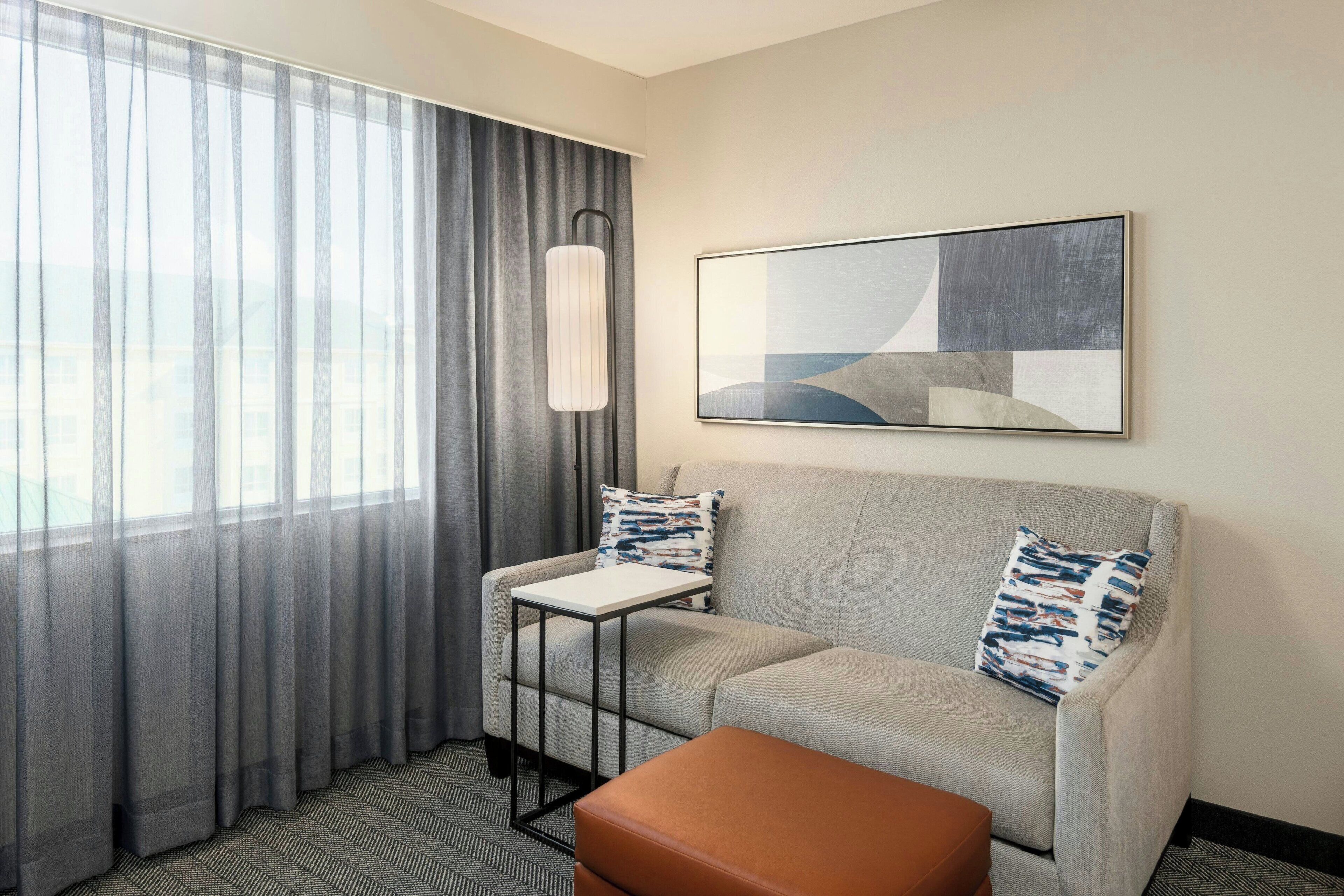 Habitación de hotel con sofá gris, una mesita, un reposapiés marrón, una lámpara de pie y un cuadro abstracto grande en la pared.