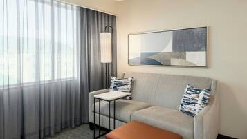 Habitación de hotel con sofá gris, una mesita, un reposapiés marrón, una lámpara de pie y un cuadro abstracto grande en la pared.