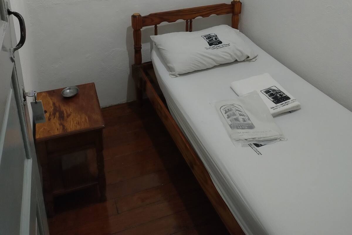 Una habitación pequeña con una cama individual, cabecera y mesita de noche de madera, y un cuenco metálico en el suelo.