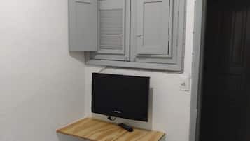 Quarto com cama, televisão na parede, prateleira de madeira e janela com persianas.