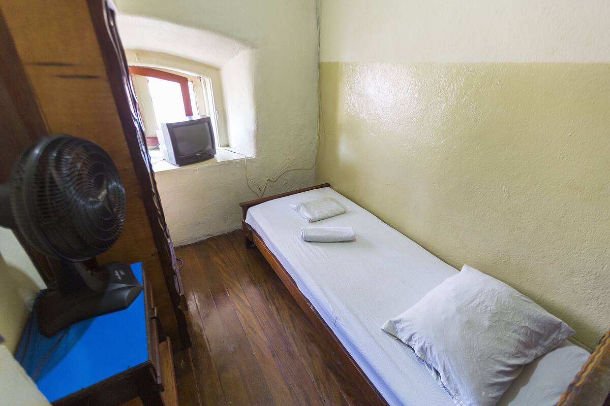 Una habitación pequeña con cama individual, piso de madera, un escritorio con computadora y un ventilador de pie.