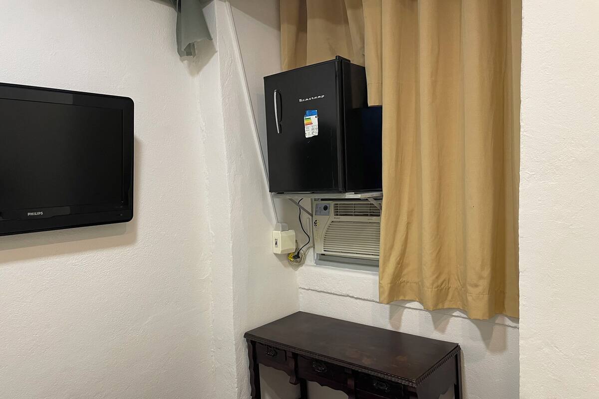 Una habitación con televisor de pantalla plana montado en la pared, una mesita de madera y una ventana con cortinas color beige.
