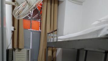 Quarto compacto com uma cama beliche, uma mesinha com um controle remoto, um aquecedor e uma janela com cortinas.