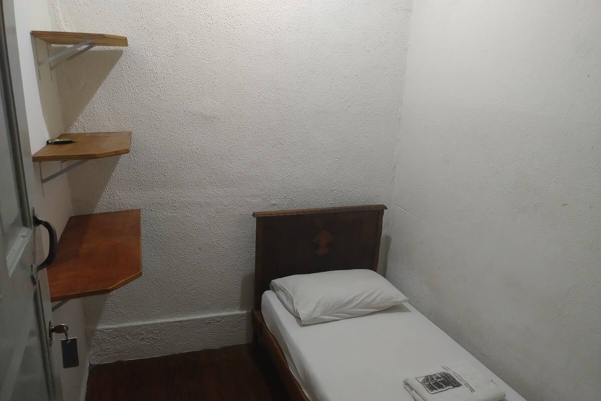 Una habitación pequeña con una cama individual, cabecera y estante de madera, además de una mesita.