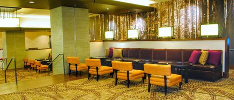 Un hall de hotel con alfombra estampada, una zona de estar con sillas anaranjadas, un sofá con cojines y una pared con persianas verticales.