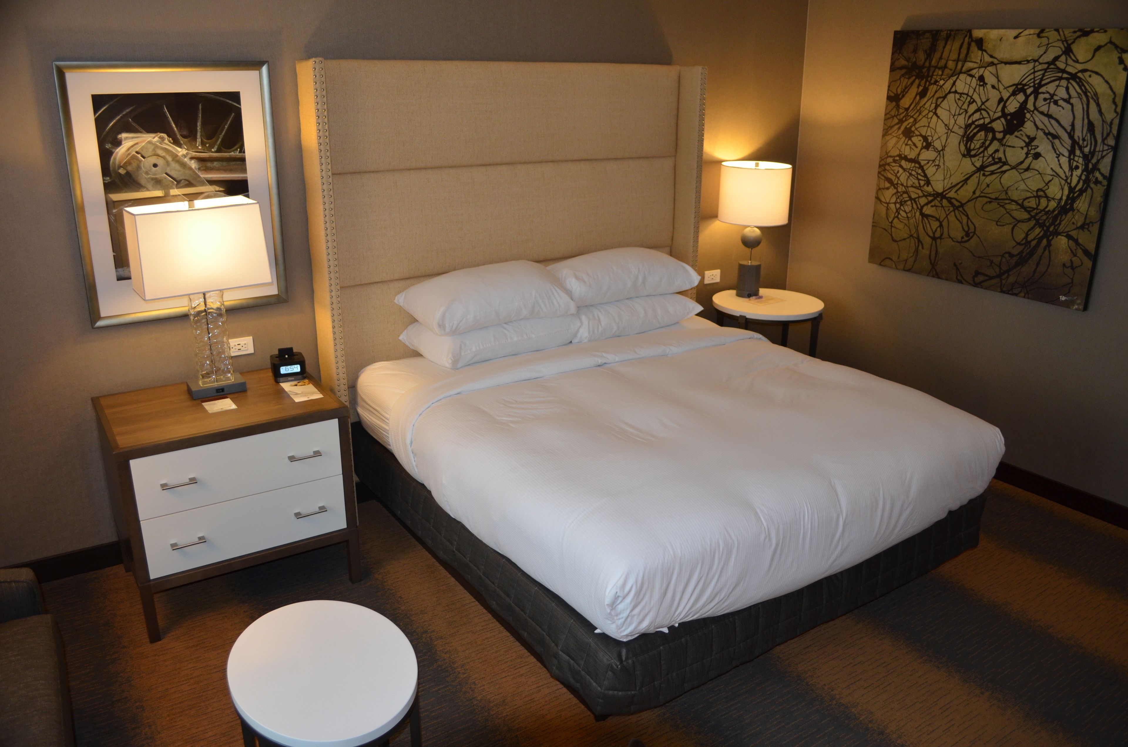 A neatly made bed with white linens, a bedside table with a lamp, a small round table, and a wall-mounted artwork.