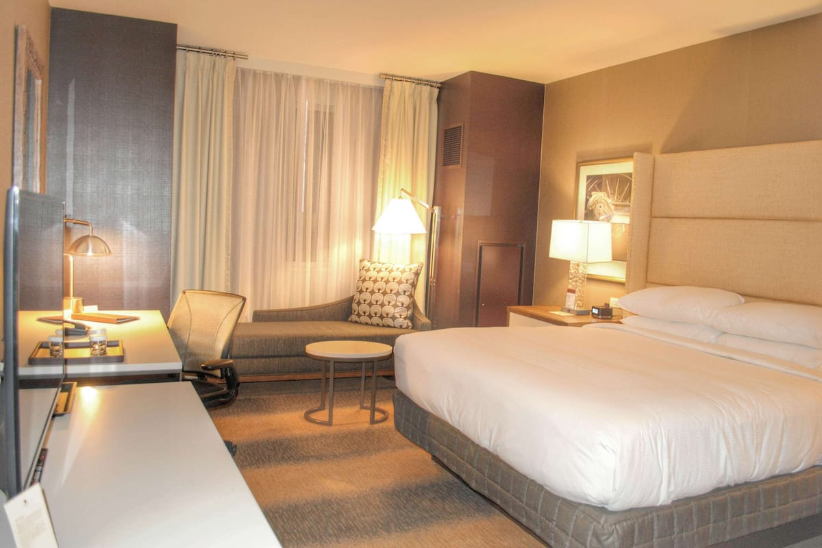 A hotel room with a bed, a desk, a chair, a small table, and a lamp.