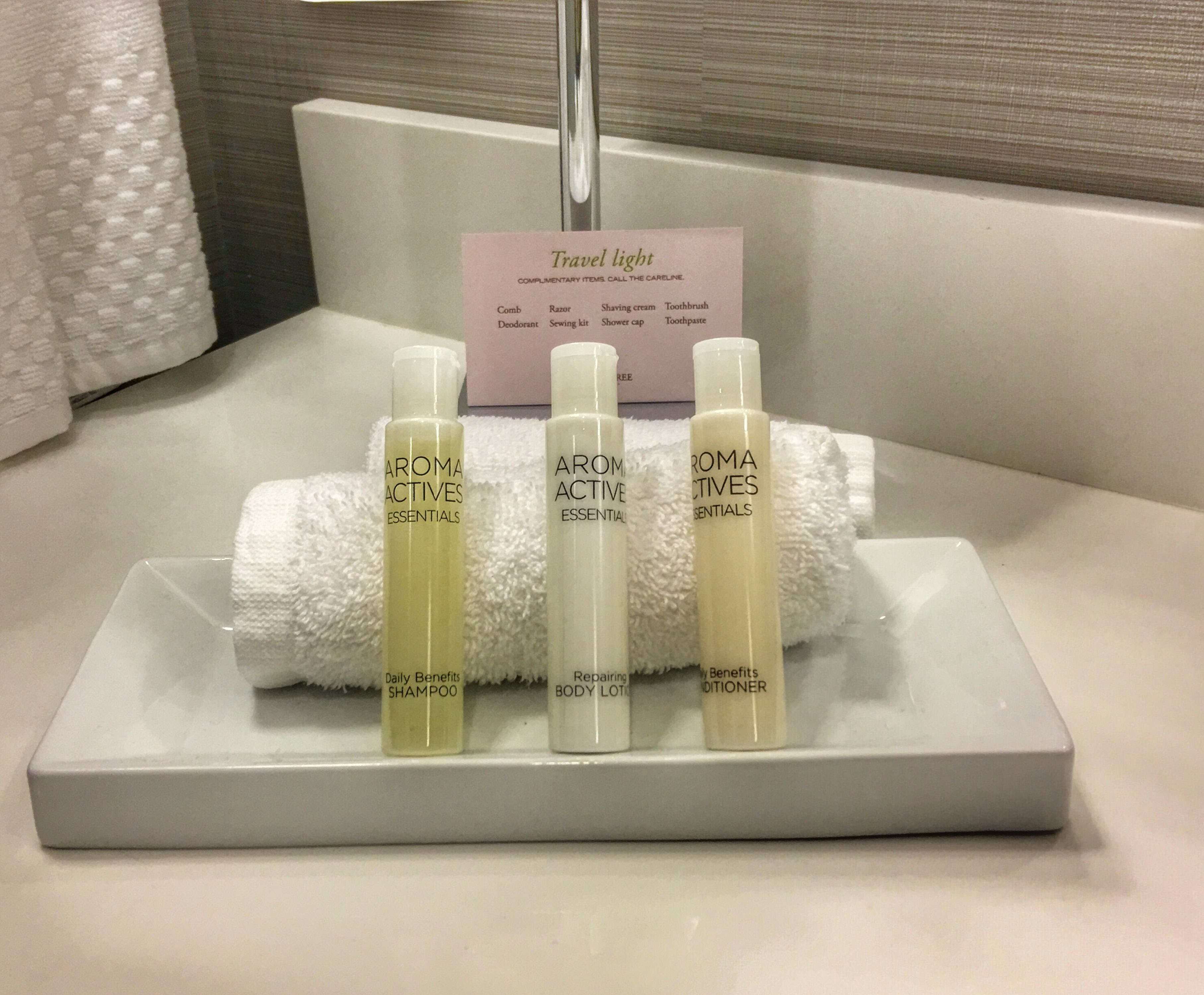 Un tocador de baño con un lavabo blanco, una toalla y tres botellas de productos 'AROMA ACTIVE ESSENTIALS': 'Champú Beneficios Diarios', 'Loción Corporal Reparadora' y 'Acondicionador Beneficios'. Se observa una tarjeta con la inscripción 'Travel light' al fondo.