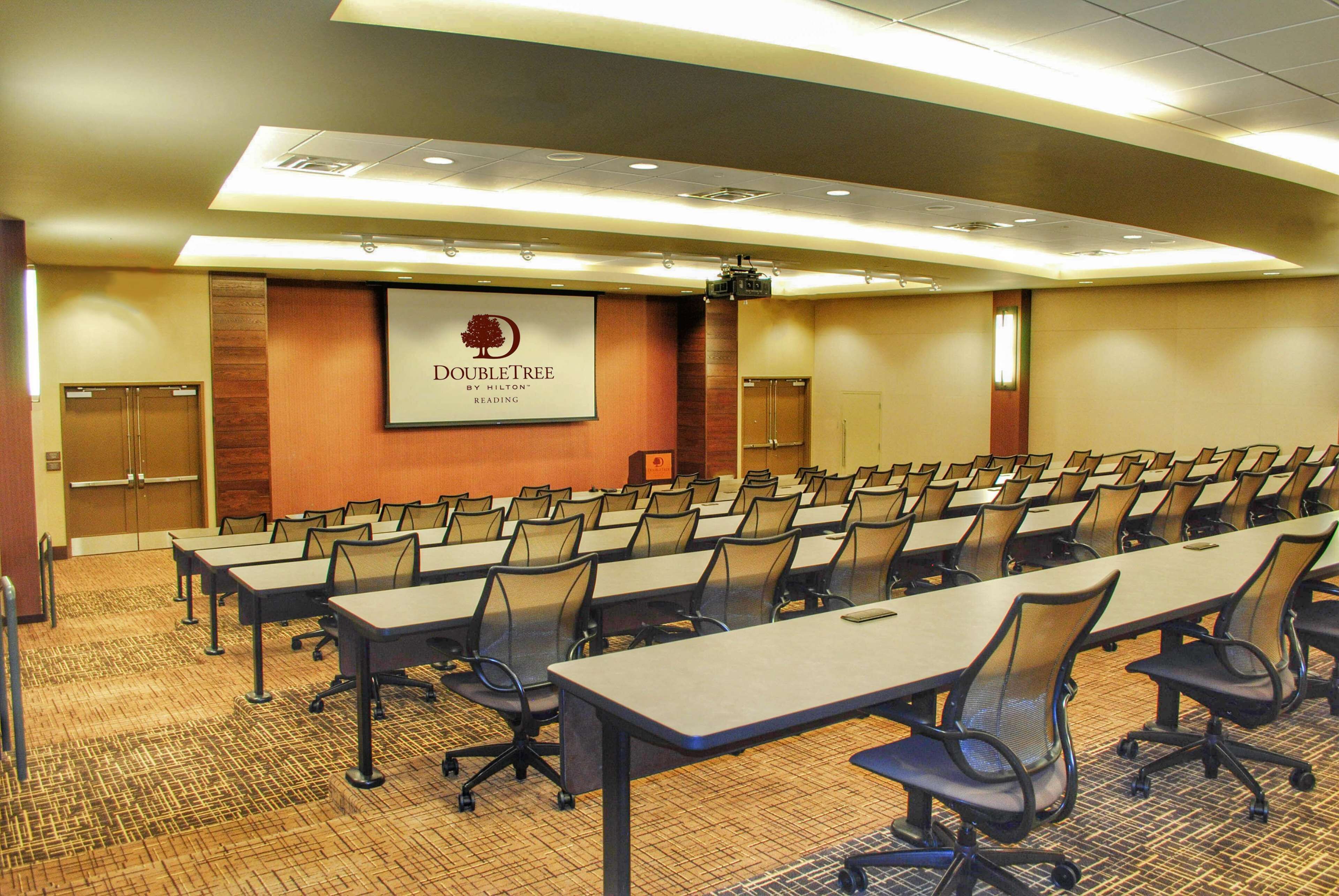 Un salón de conferencias con filas de mesas y sillas, una pantalla de proyector y un cartel que dice "DOUBLETREE by Hilton Reading".