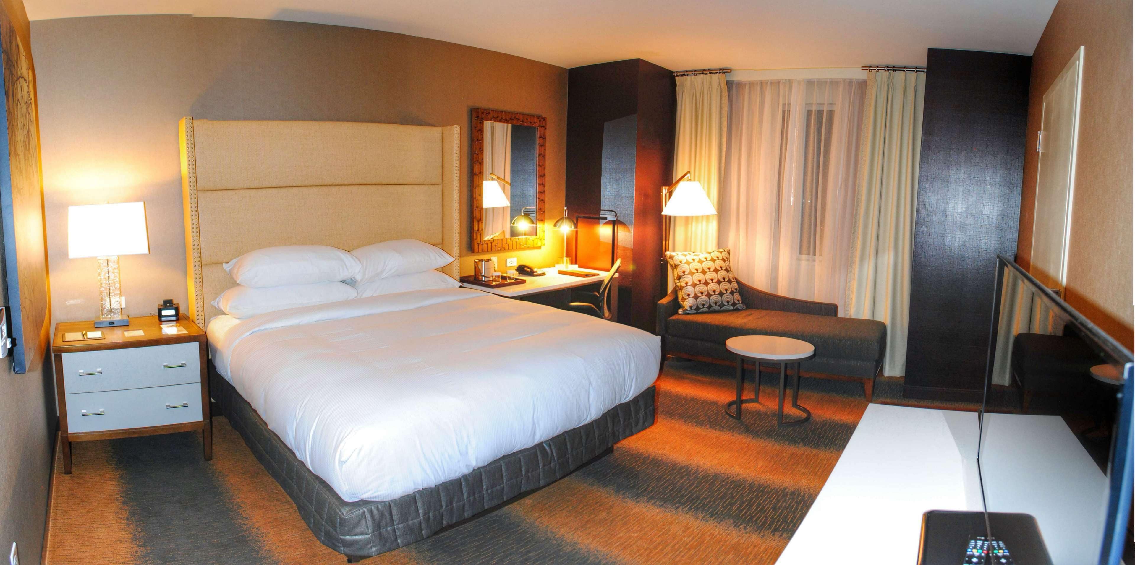 A hotel room with a large bed, a desk, a chair, a small table, and a TV.
