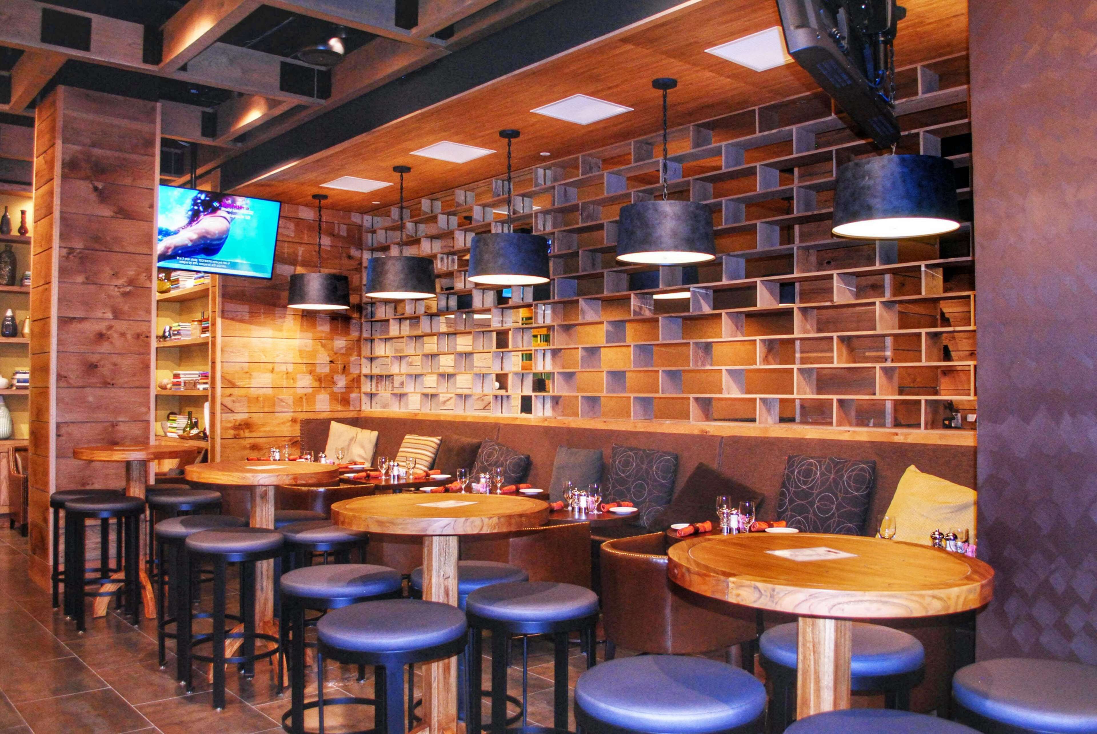 Un espace bar avec plusieurs tables et tabourets en bois, des étagères remplies de bouteilles et un téléviseur fixé au mur.
