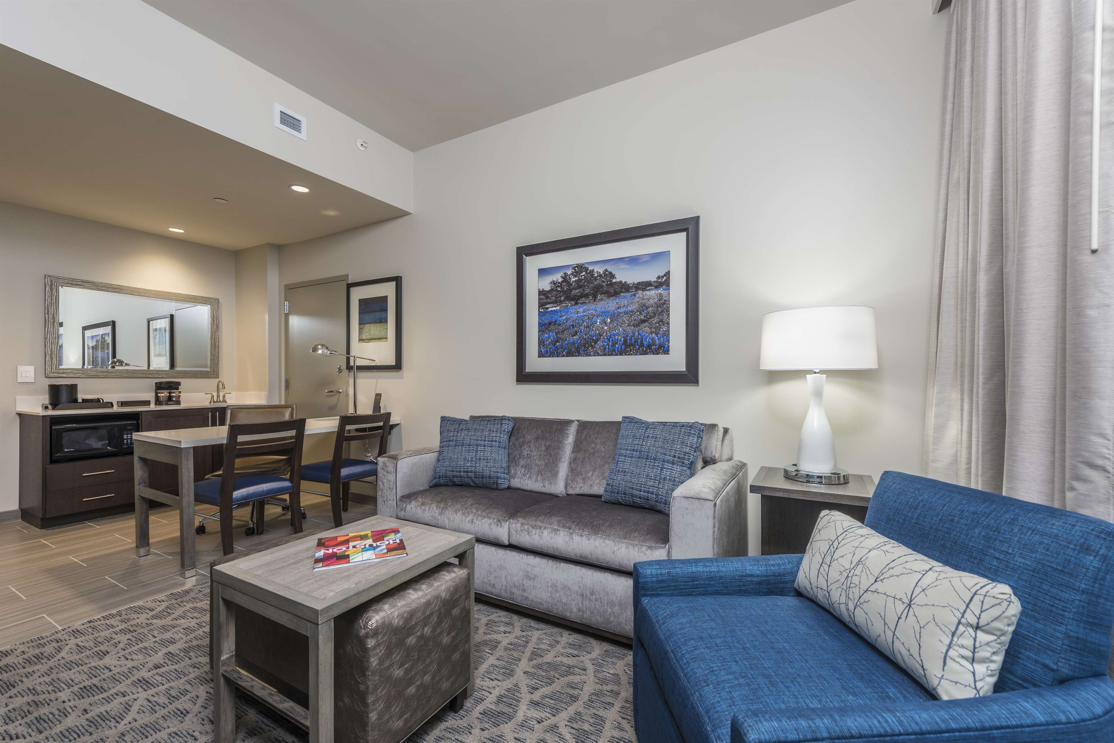 A modern hotel room with a grey sofa, a blue sofa, a dining area with a table and chairs, a small coffee table, a lamp, and a framed picture on the wall.
